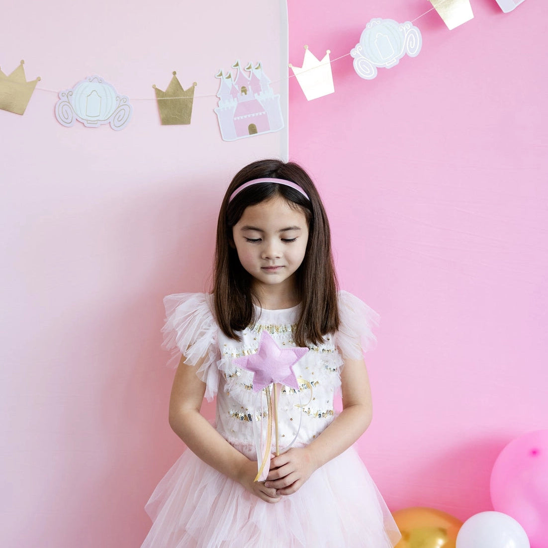 Little girl in frilly dress holding pink princess wand with eyes closed, crown-and-carriage garland behind her.