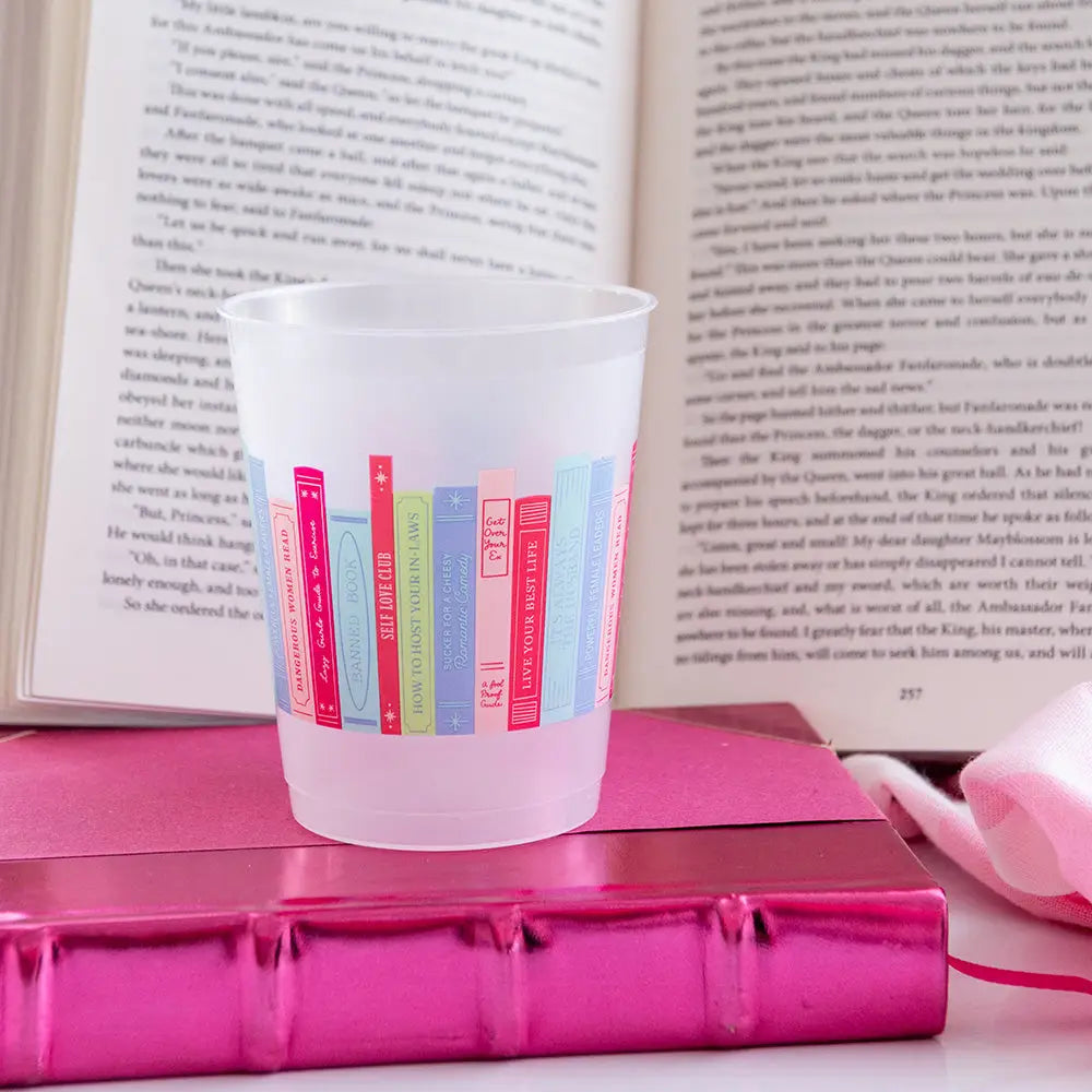 Frosted plastic cup with pastel bookshelf pattern resting on hot-pink journal, open novel in background.