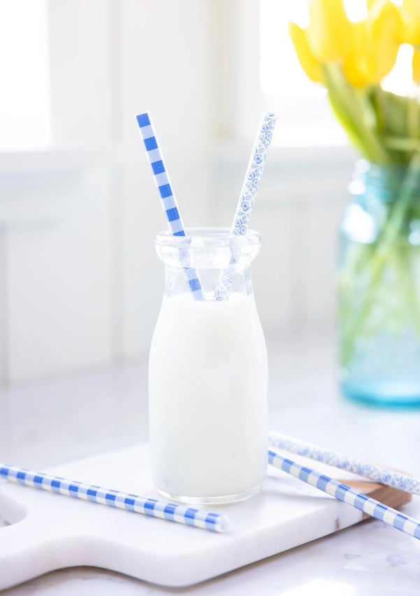 Lifestyle Scene: Two patterned blue straws in a glass milk bottle on a marble board with yellow tulips in background.