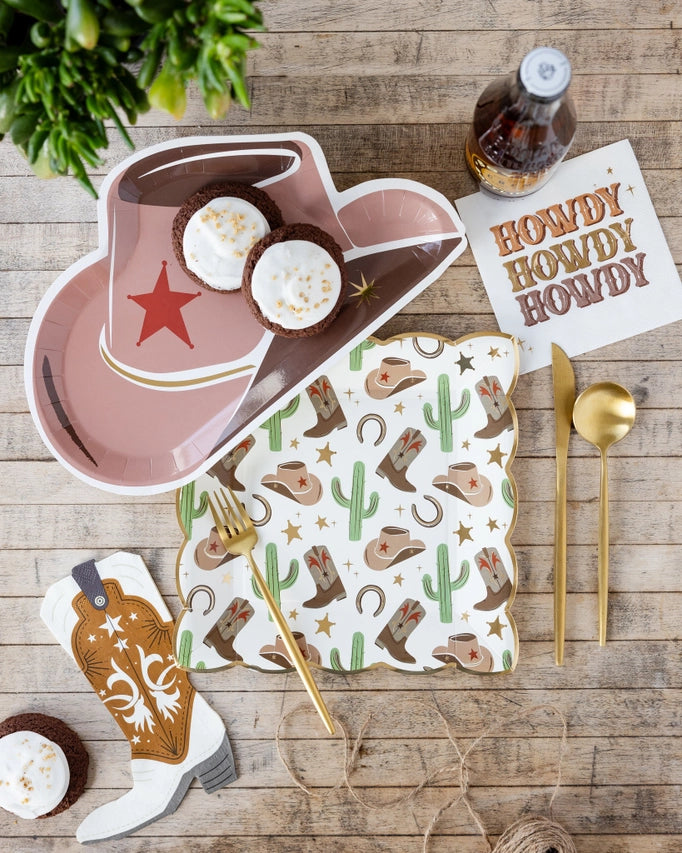 Rustic wood table set with cowboy hat plate, cactus print plates, gold cutlery, and “Howdy” napkin alongside mini cupcakes.