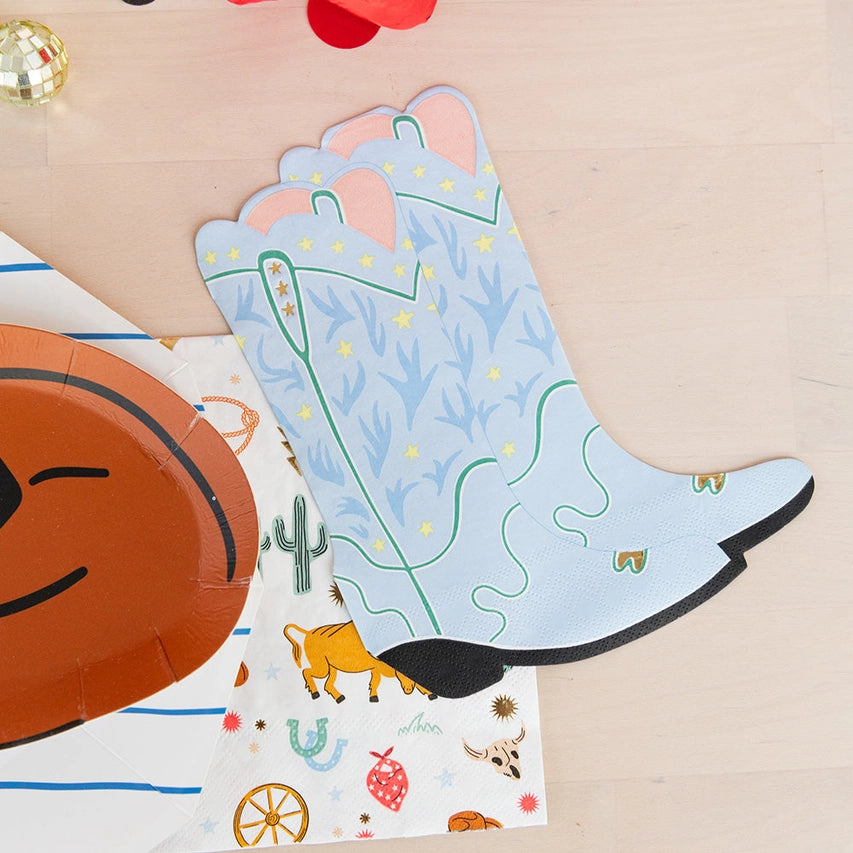 Two layered dusty‑blue boot napkins atop patterned horse napkin and beside a rust hat plate.