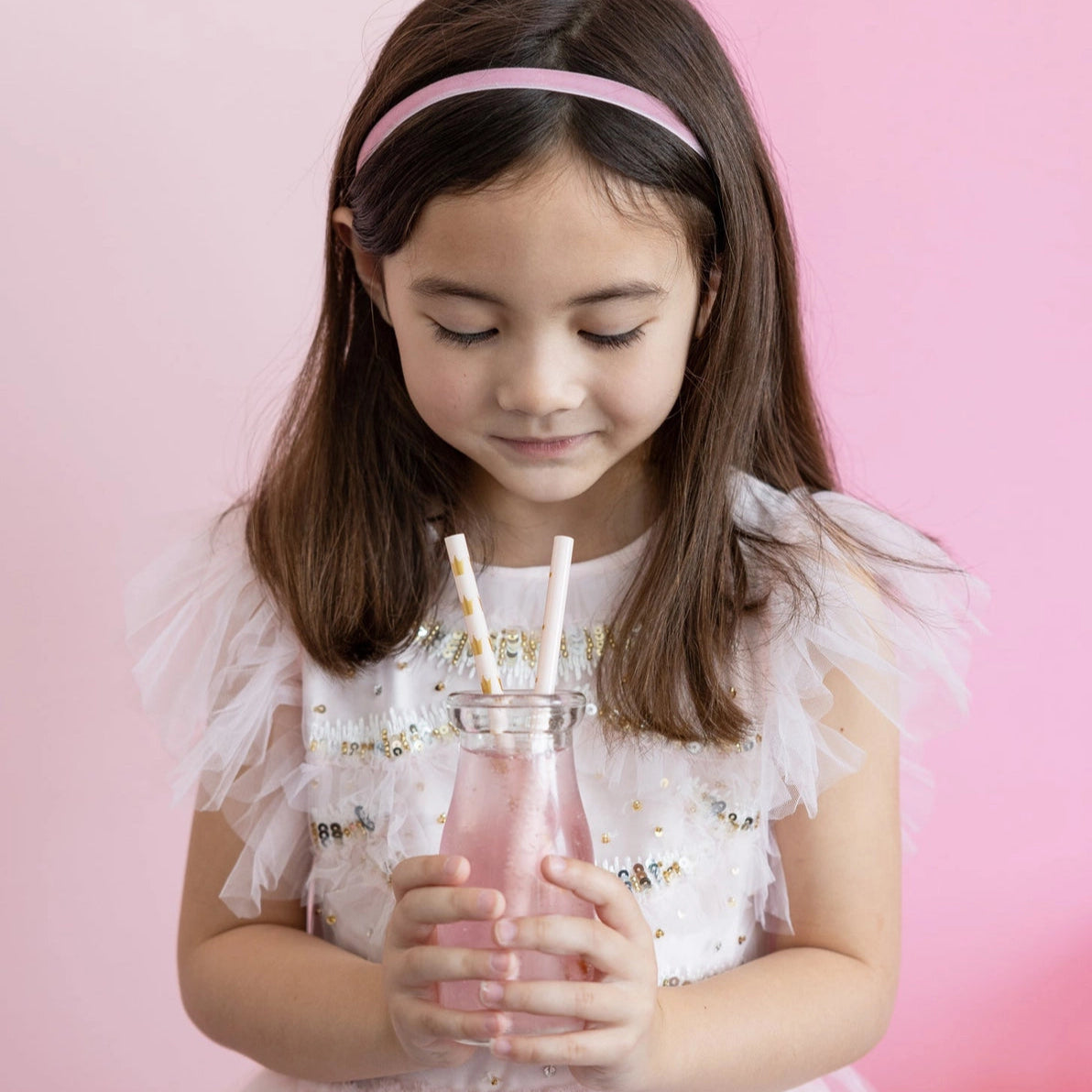 Close-up of child holding glass bottle with two pink straws against pink backdrop.