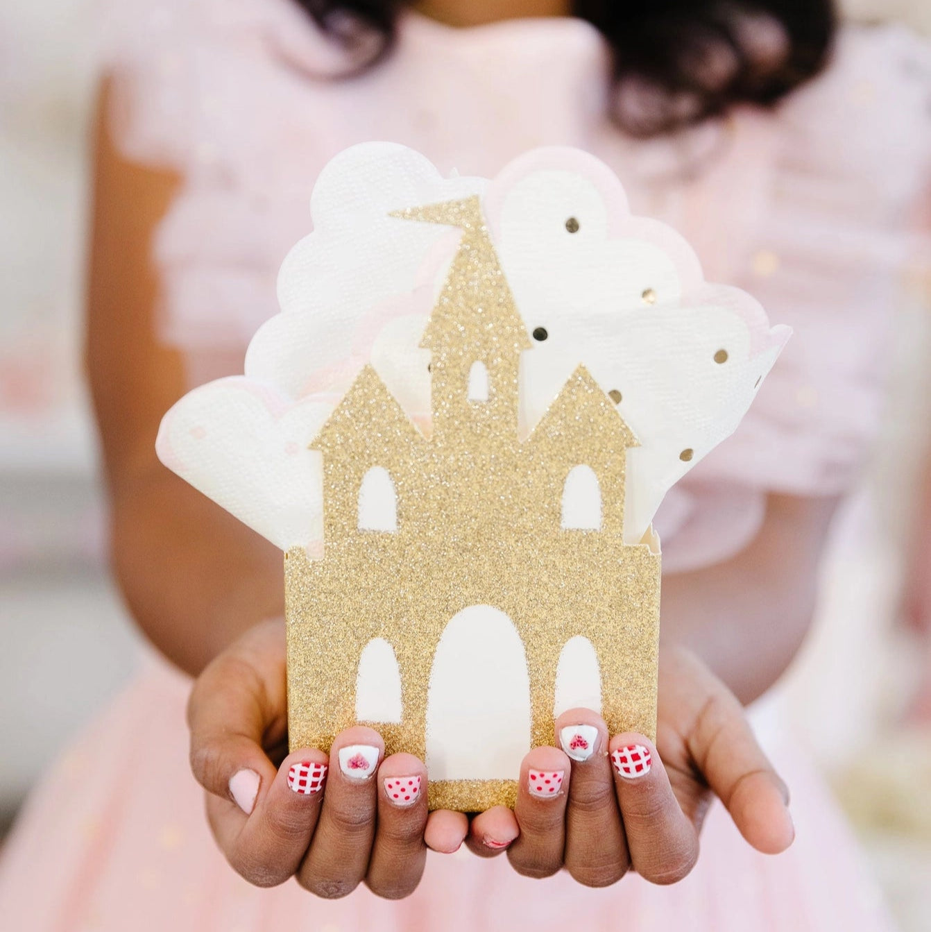 Child in pink dress presenting gold-glitter castle favor box filled with polka-dot napkins.