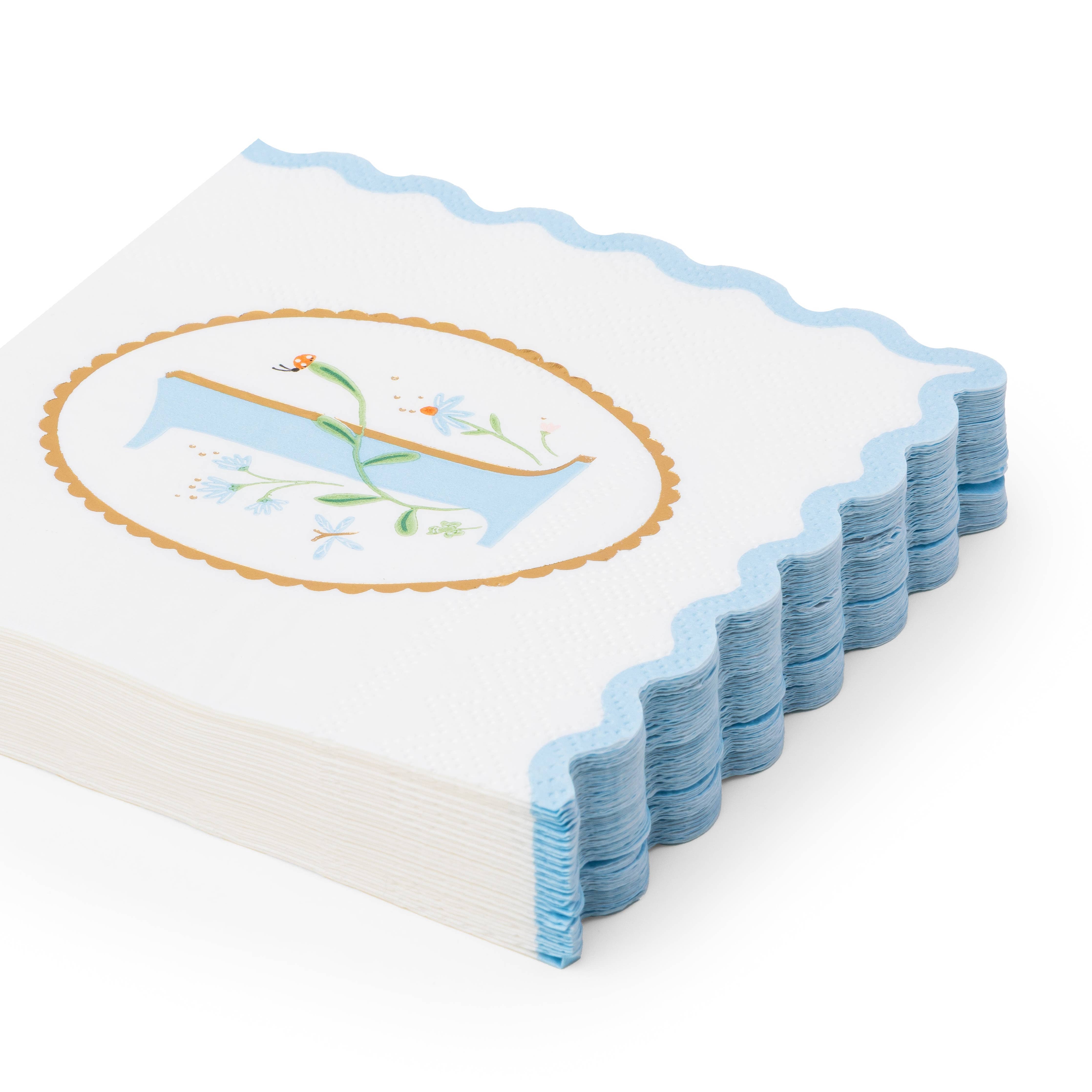 Angled stack of white first birthday boy paper napkins with sky blue floral illustrated number one in gold oval medallion and blue scalloped edge on white background
