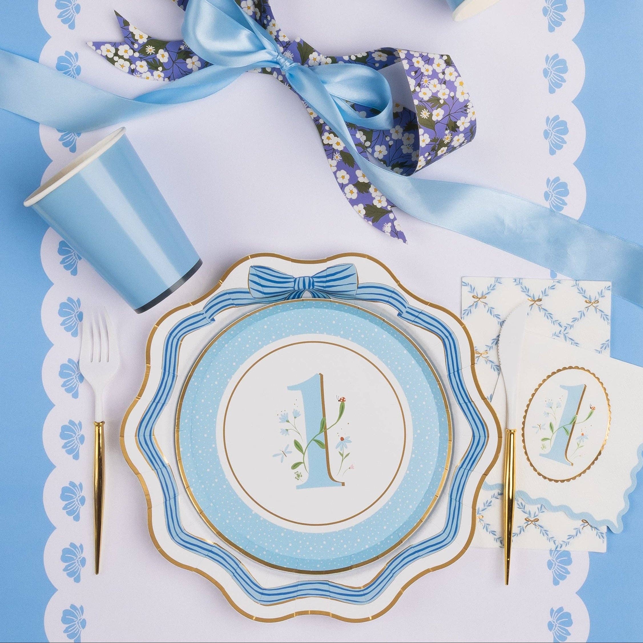 Overhead lifestyle shot of sky blue first birthday boy party table with floral number one cake plate stacked on bow dinner plate, matching napkin, blue cup, gold cutlery and blue satin ribbon on white scalloped placemat
