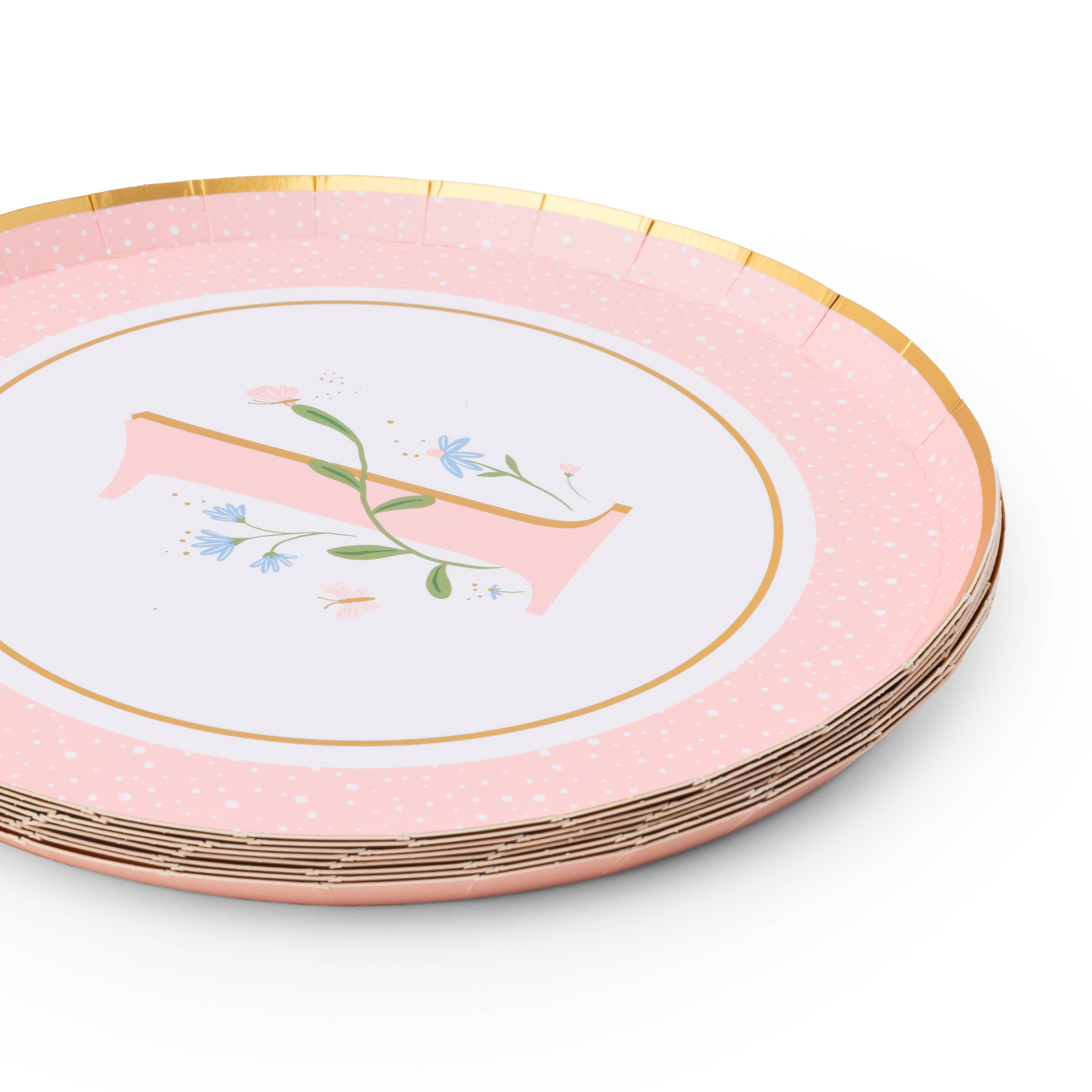 Angled stack of petal pink polka dot first birthday paper plates with floral illustrated number one and gold foil rim on white background