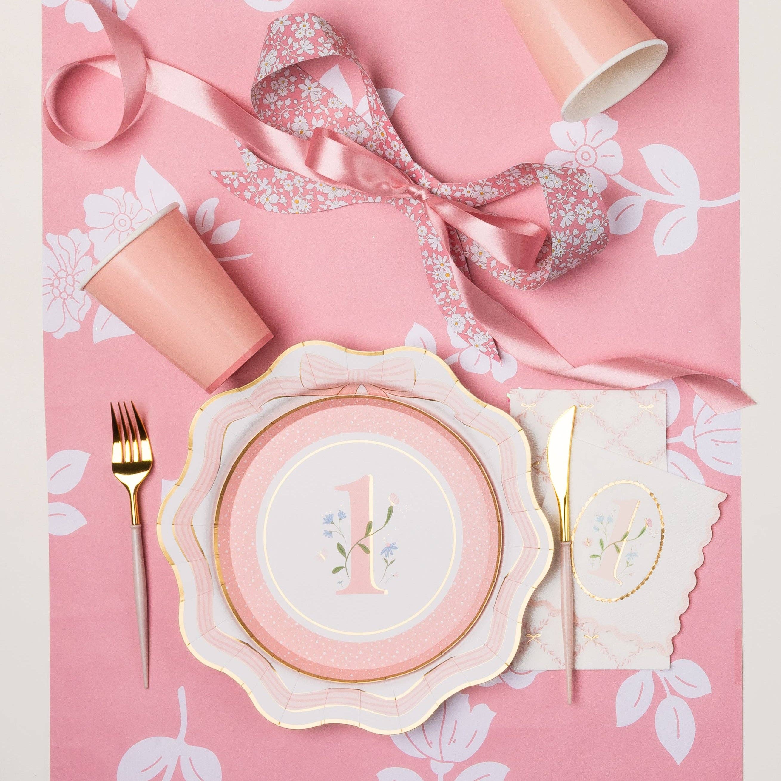 Overhead lifestyle shot of pink floral first birthday girl party table with number one cake plate stacked on scalloped dinner plate, matching napkin, pink cup, gold cutlery and satin ribbon on pink floral tablecloth
