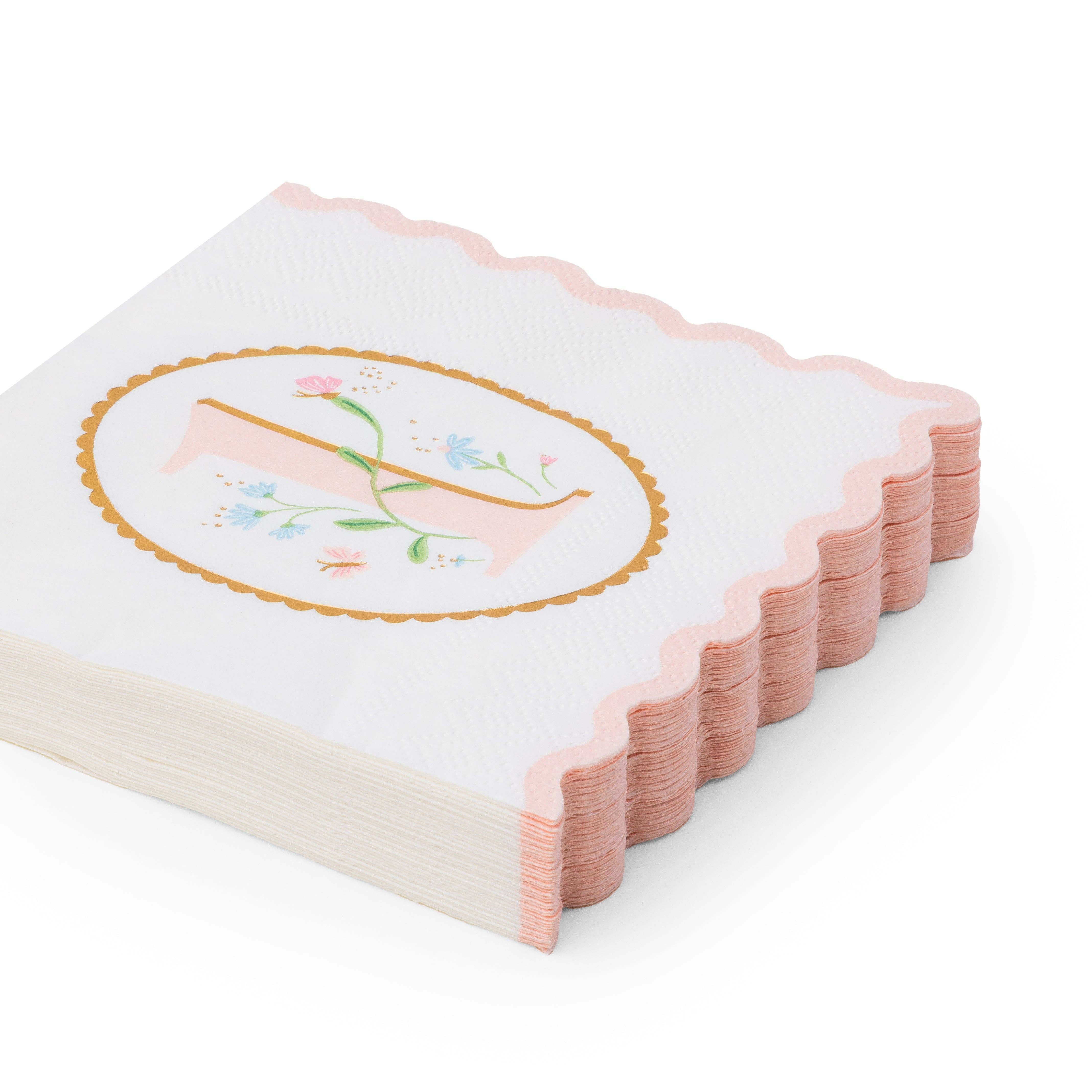 Angled stack of white 1st birthday girl paper napkins with pink floral illustrated number one in gold oval frame and pink scalloped edge on white background