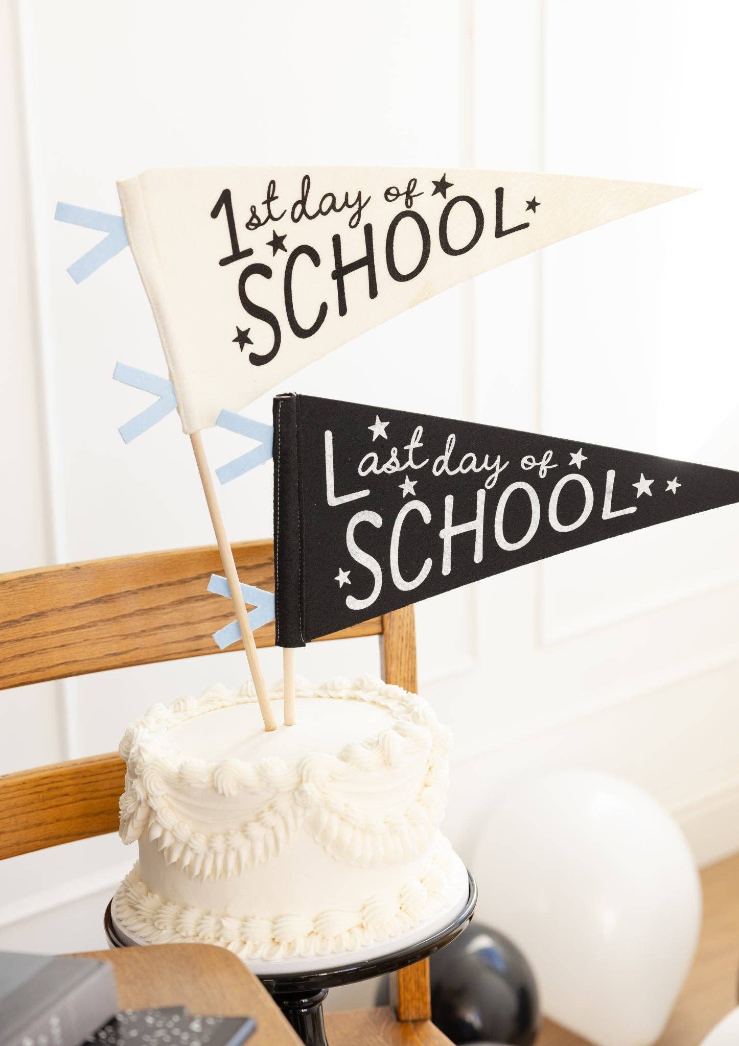 Close-up of 1st Day and Last Day of School felt pennants used as cake toppers on a white layer cake with white panel wall and black balloon background