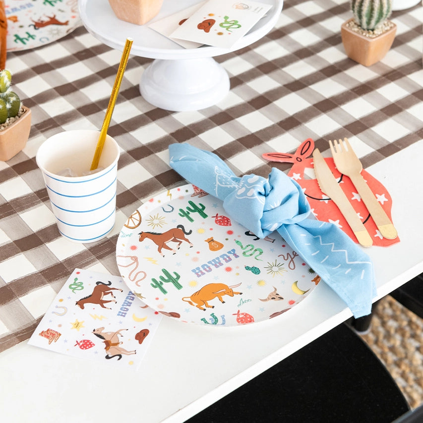 Place setting close‑up (blue napkin): Close view of a single cowboy‑pattern plate with blue bandana napkin, striped paper cup, and gingham tablecloth.