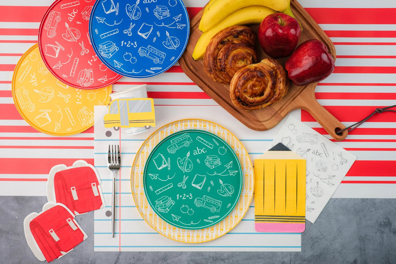 Colorful brunch setup with red, blue, and yellow doodle plates, pastries, bananas, and apples arranged over red‑striped table runners and a notebook placemat.