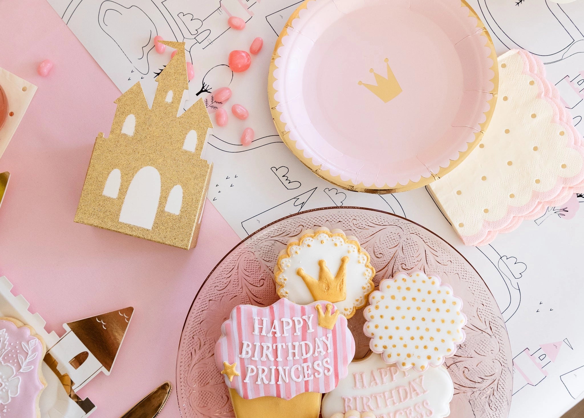 Flat-lay of party table: glitter castle box, crown plate, royal cookies, and pink candies on map-style placemat.