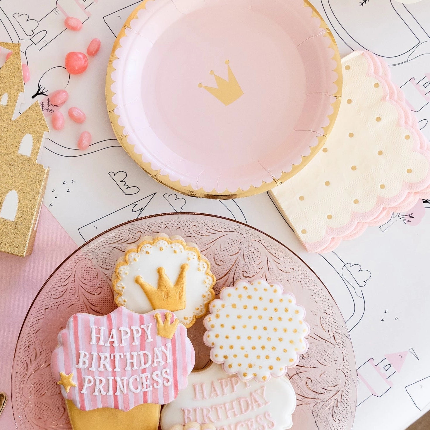 Flat-lay of gold castle favor box, blush crown plate, and scalloped polka-dot napkin arranged on pink table for a princess birthday party.