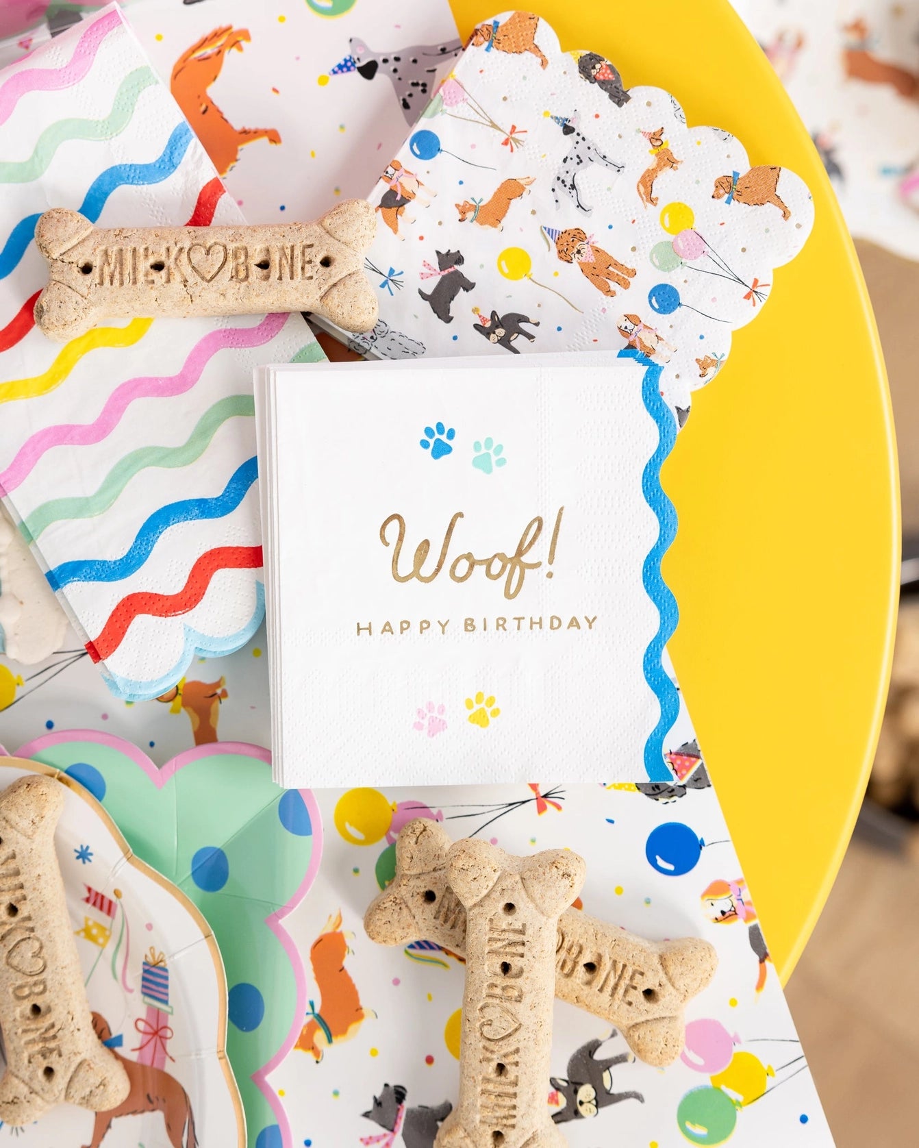 Party close-up: rainbow napkin, dog-print napkin, and “Woof! Happy Birthday” cocktail napkin layered on a bright yellow table with bone-shaped treats scattered around.