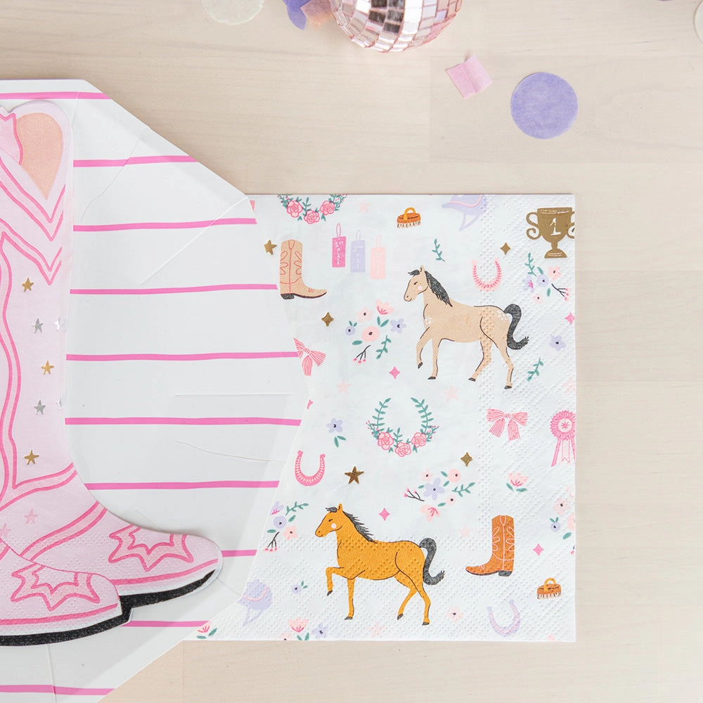 A pink-striped octagon plate topped with a die-cut cowgirl boot napkin, next to a square horse-print napkin featuring tan horses, floral wreaths, and ribbons; scattered confetti and shiny disco balls decorate the pastel tabletop.