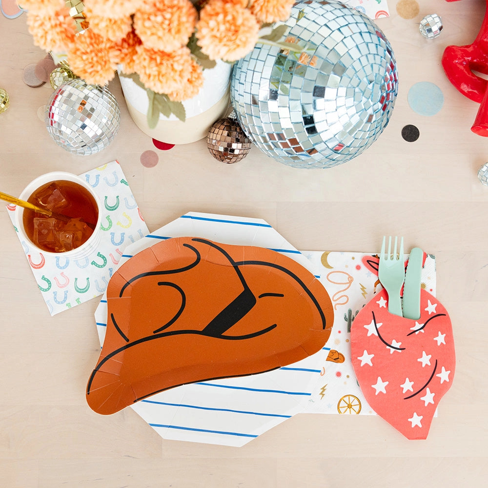 Close‑up setting: Brown cowboy‑hat plate on a blue‑striped charger next to a white napkin covered in tiny multicolor horseshoes beside a cup of iced tea.