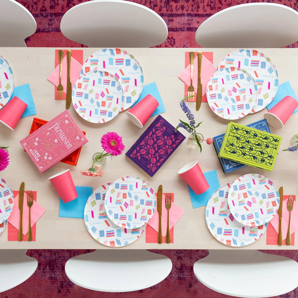 Overhead tablescape with colorful book-pattern plates, pink cups, gold cutlery, and classic novels as centerpieces.
