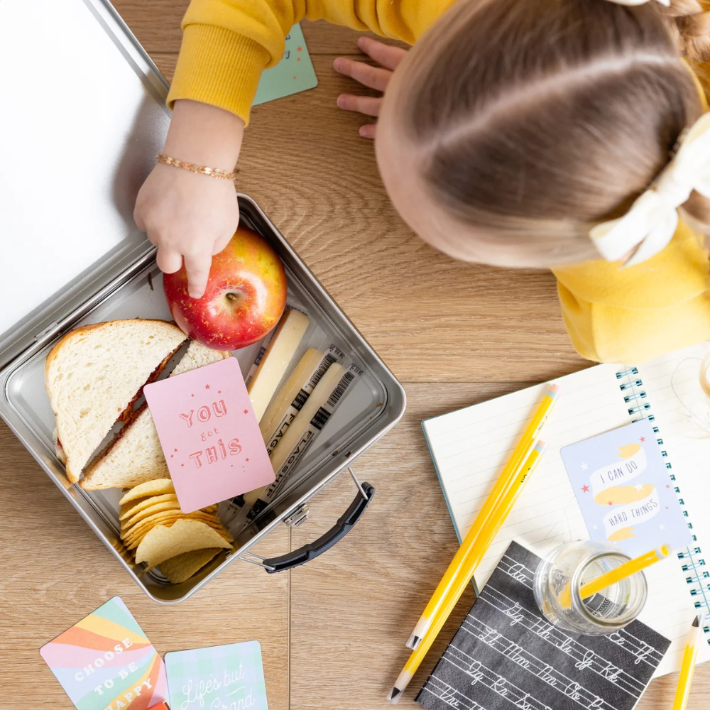 Overhead view of a child placing a “You Got This” card into a lunchbox.