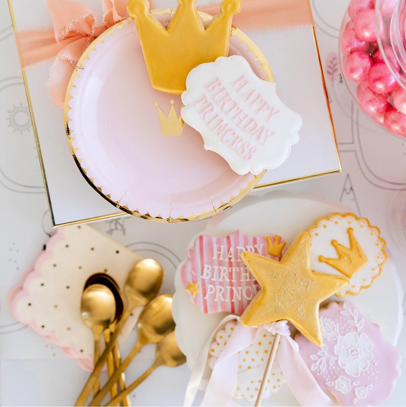 Gift-box vignette featuring stack of pink crown plates and ribbon, with gold spoons resting on scalloped polka-dot napkins—perfect baby girl baby shower setup.