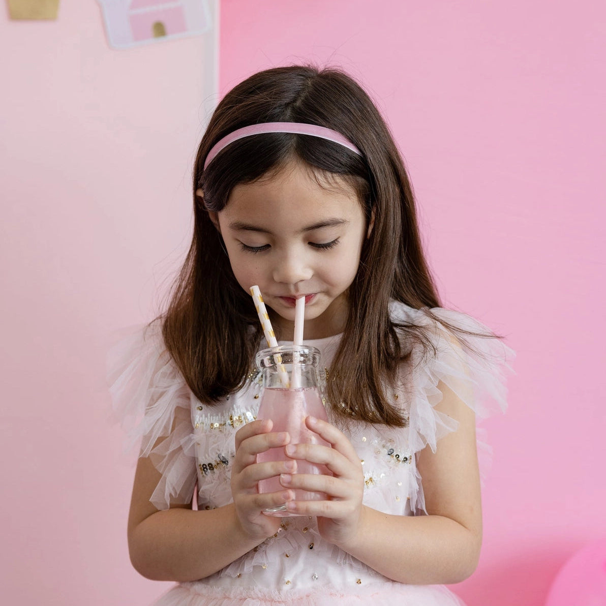 Little girl sipping pink drink through two crown-pattern pink straws at princess party.