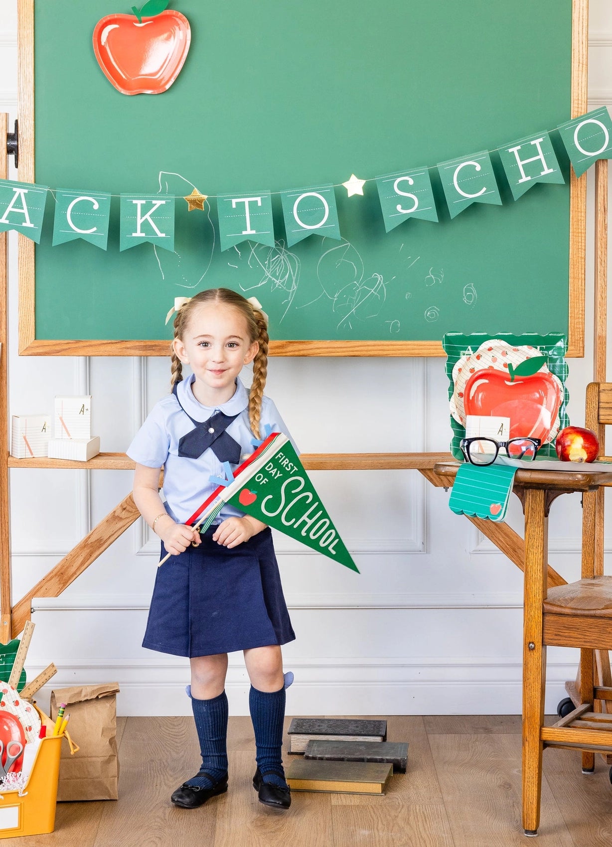 A young girl standing in front of a green chalkboard decorated for a back to school party, with green paper napkin on a desk.