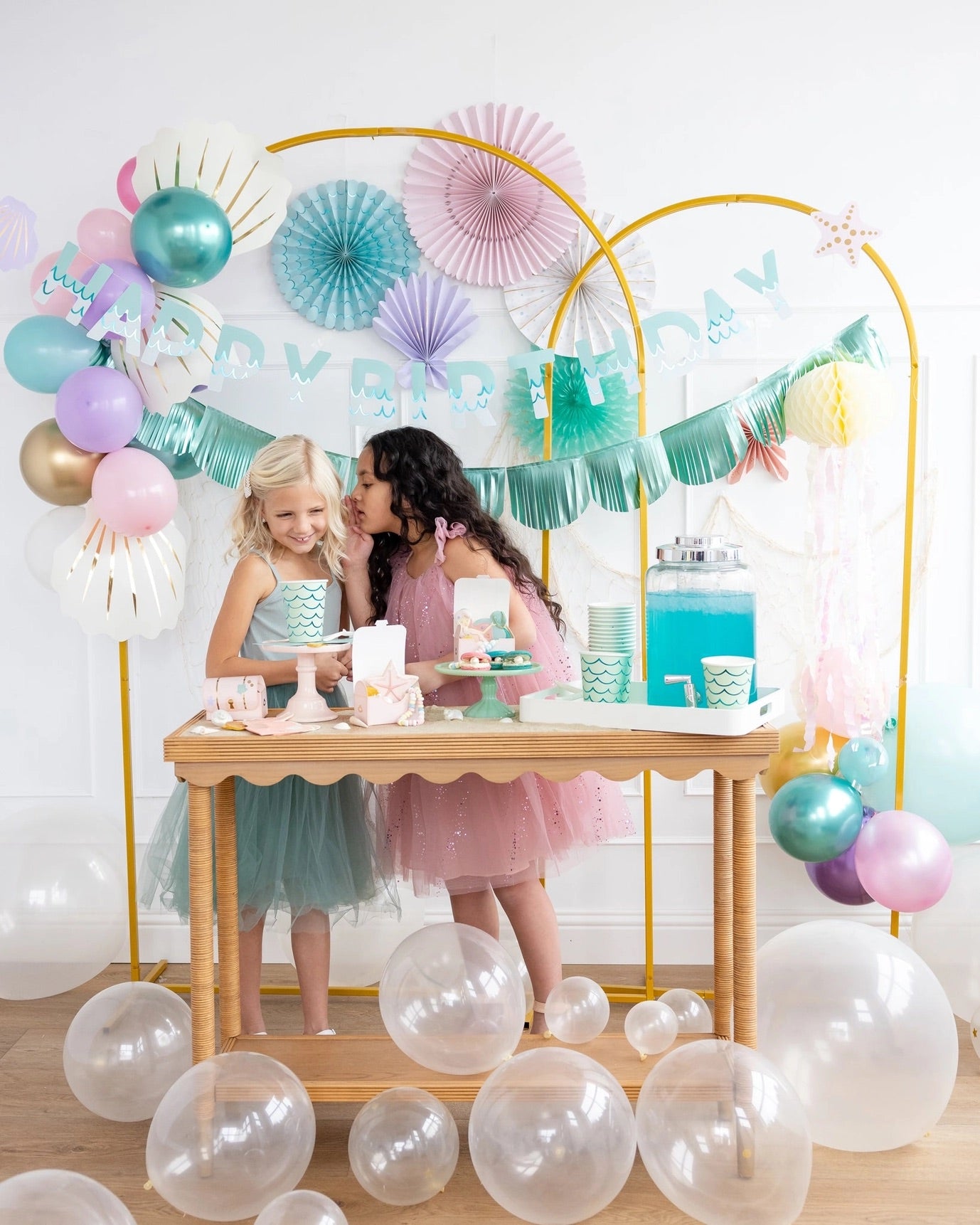 Two young girls whispering and giggling together at a festive mermaid-themed dessert table surrounded by balloons and pastel décor.