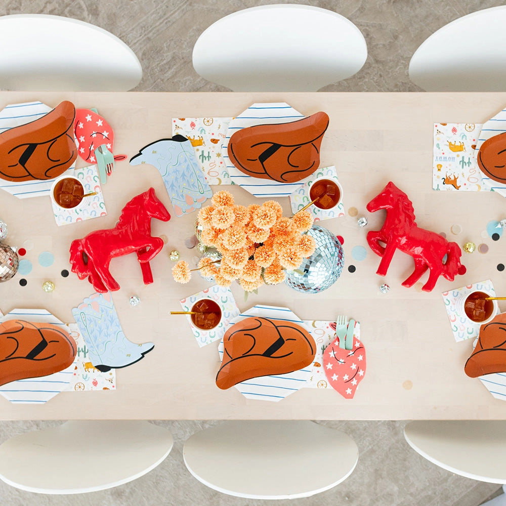 Overhead tablescape: Top‑down view of six place settings featuring brown hat plates, rainbow horseshoe napkins, blue boot napkins, red bandana cutlery pockets, orange flowers, and mirrored disco balls.