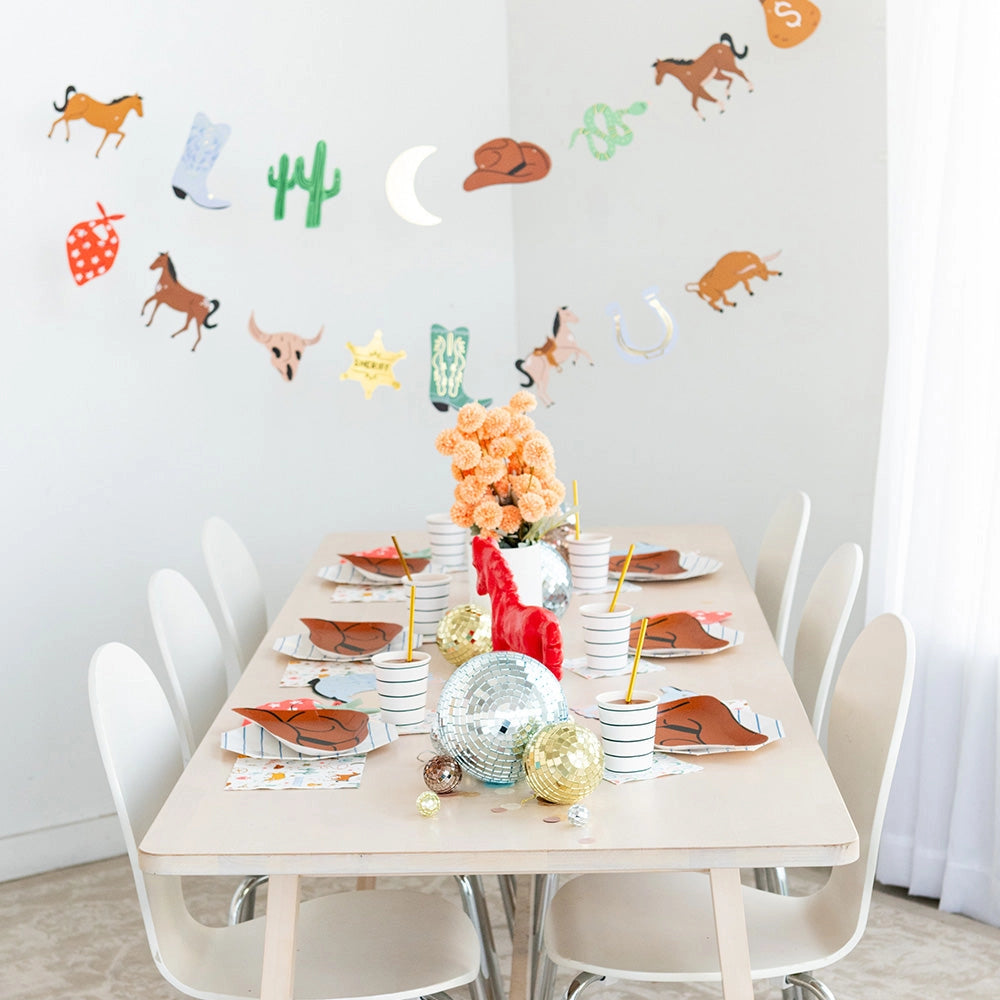 Dining‑room shot: Long blond‑wood table set for a kids’ western party with cowboy‑hat plates, striped cups, disco‑ball centerpieces, and a wall garland of cowboy icons.