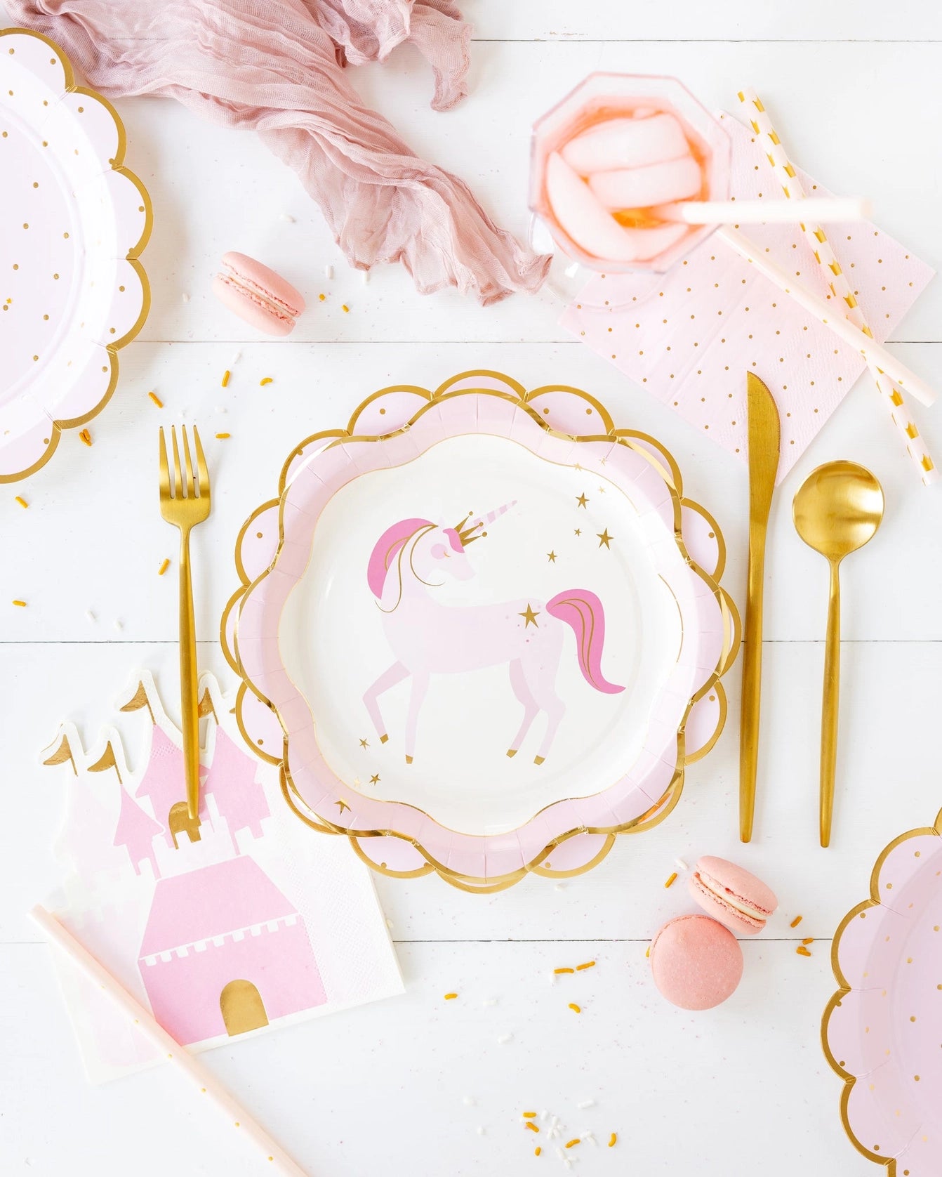 Styled dessert table: unicorn plate, castle napkin, scattered macarons, and folded pink napkins with gold foil dots.