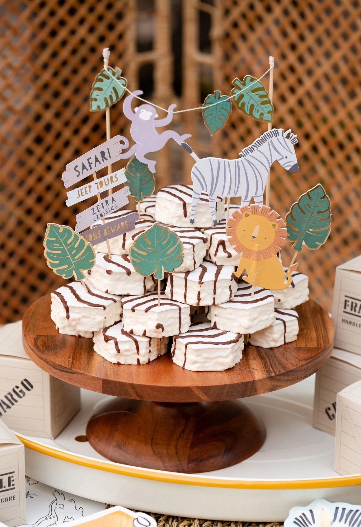 Bright, styled photo of safari animal cake toppers, including playful monkey, zebra, lion, and greenery, placed on cake bites on a wooden cake stand indoors.