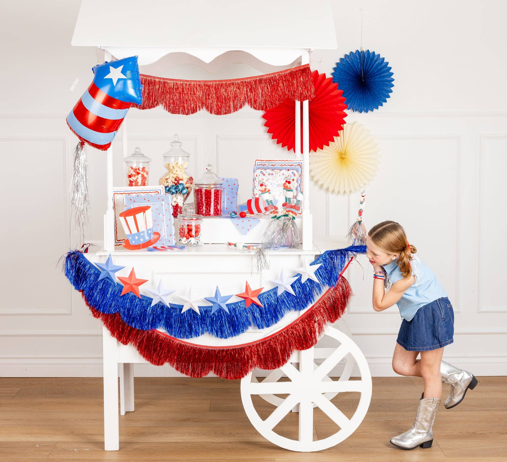 Girl in silver cowboy boots leaning on a white party cart decorated with a patriotic 3D paper star banner, red and blue fringe banners, and 4th of July tableware and candy jars