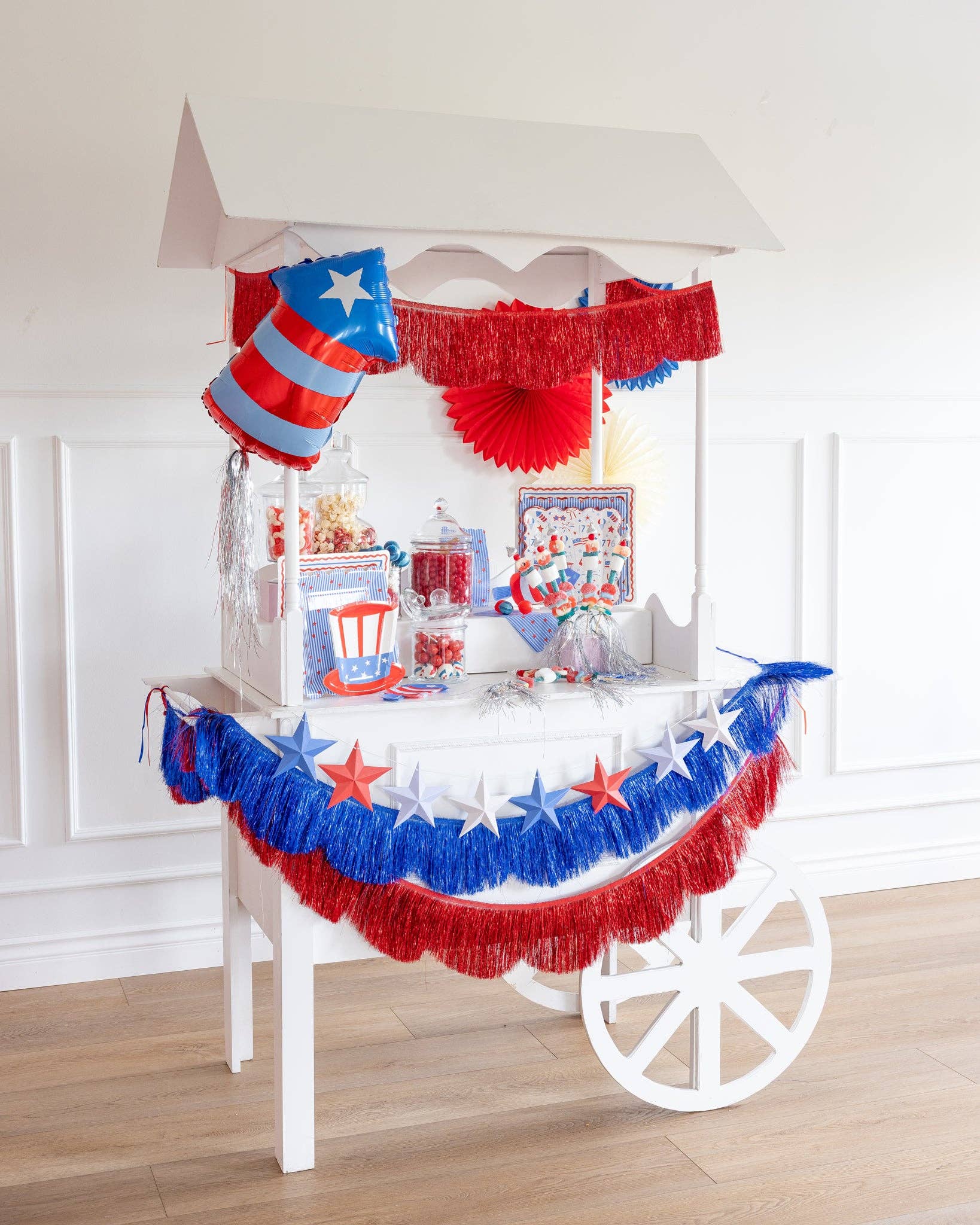 White party cart decorated for 4th of July with layered blue and red metallic fringe banners, 3D star garland, firecracker shaped balloon, paper fans, and patriotic party supplies