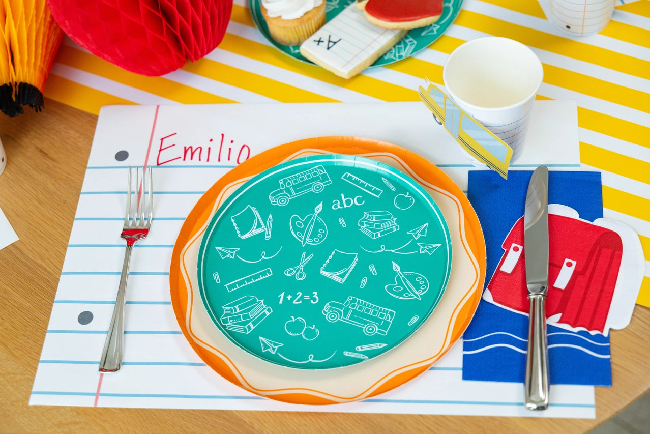 Child’s place setting labeled “Emilio” on a notebook placemat, featuring teal doodle plate, orange rim charger, and red backpack napkins beside silverware.