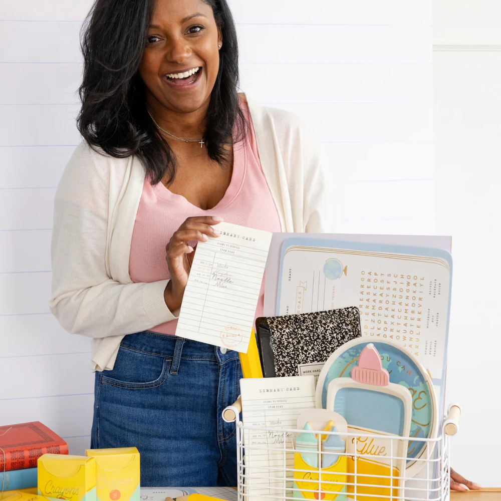 Woman smiling and holding a library card dinner napkin next to a basket filled with school-themed party supplies.