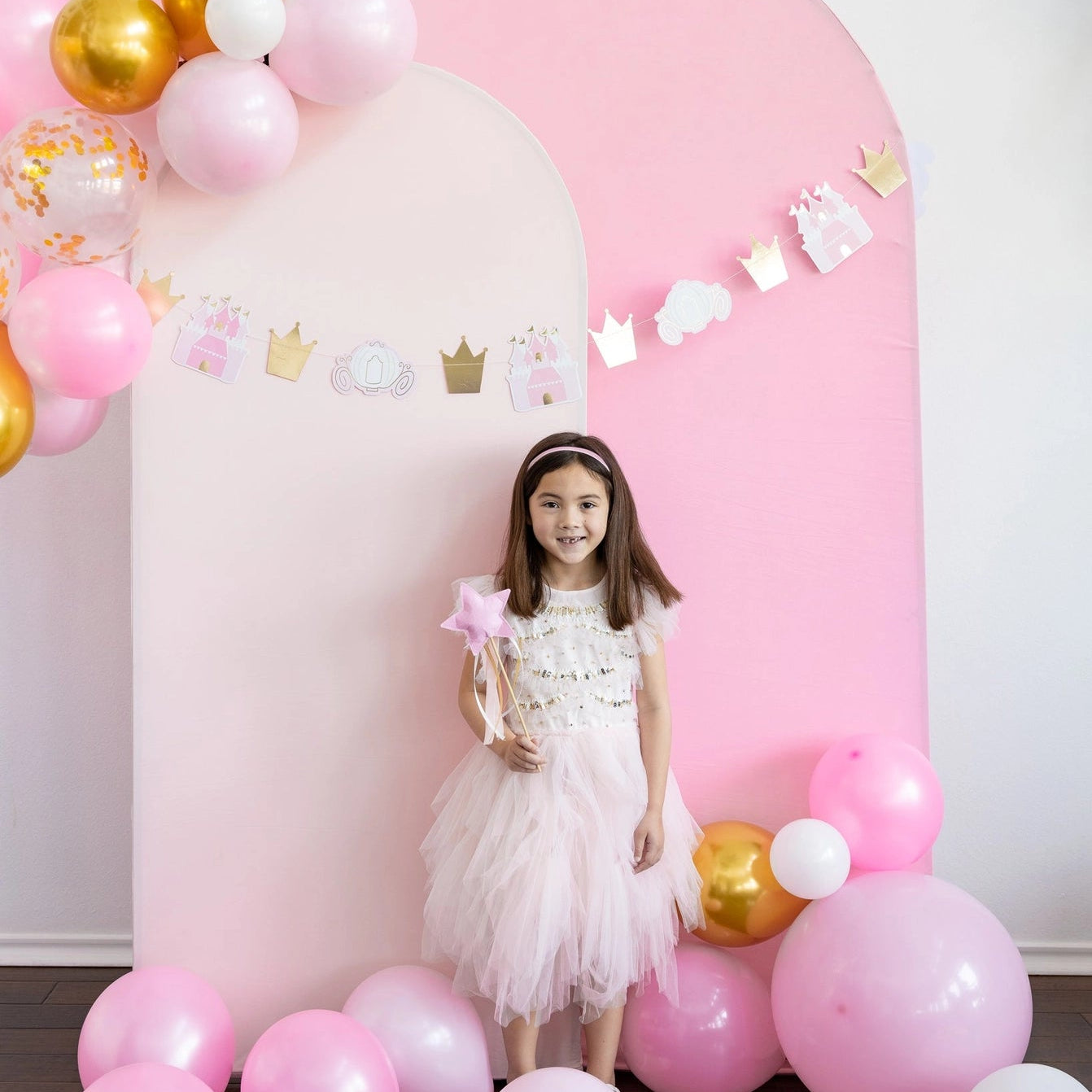 Wide shot of balloon arch and castle garland; girl in tulle dress holds princess wand at center.