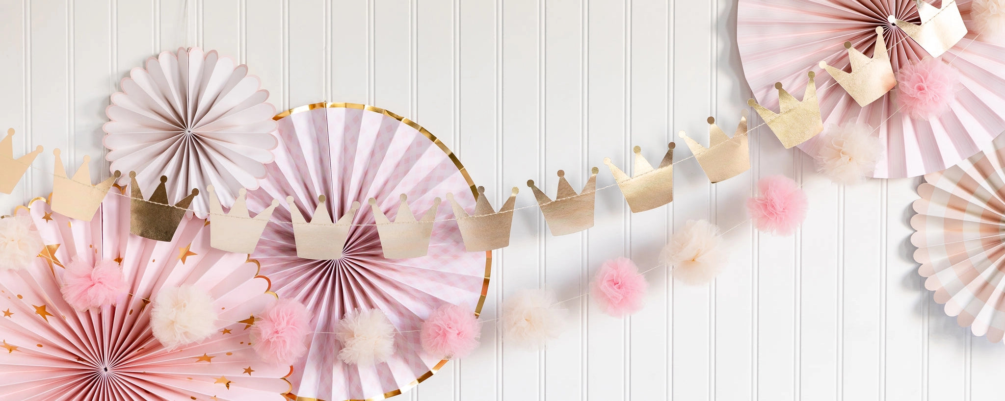 Close-up of metallic crown garland layered over pink rosette fans and tulle pom strand against beadboard wall.