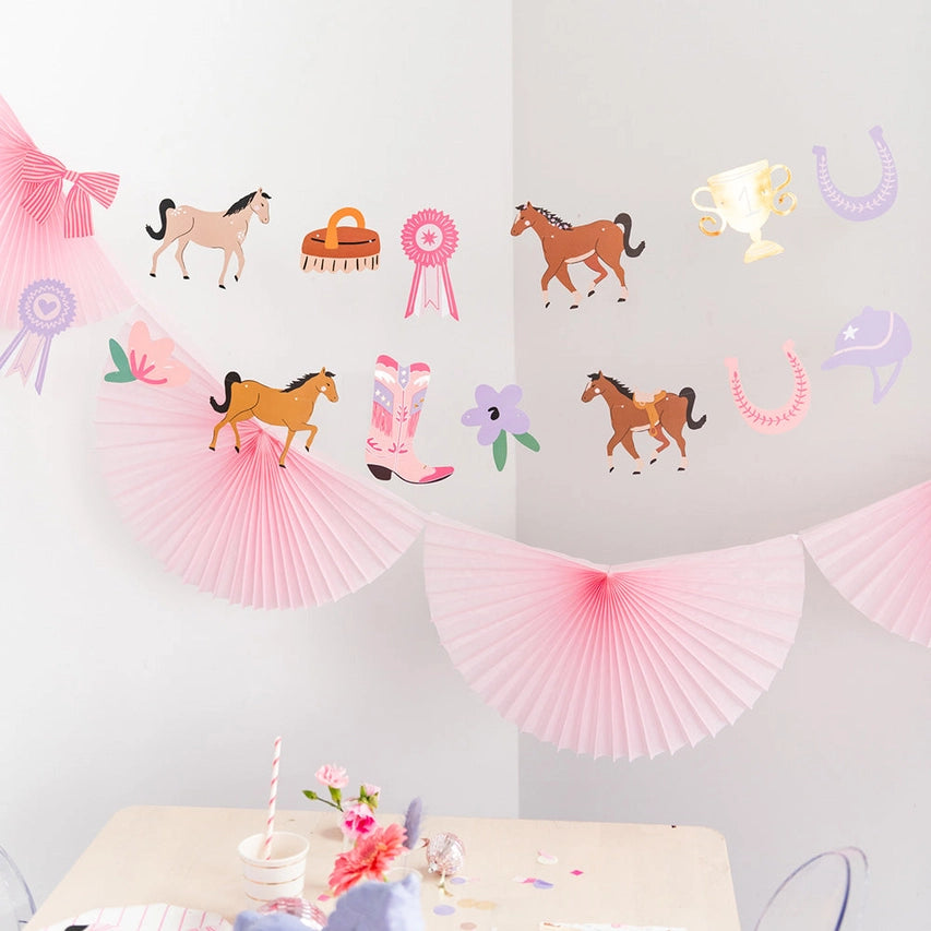  Pastel die‑cut horses, boots, ribbons, trophies, and horseshoes strung on twine above pink fan bunting.