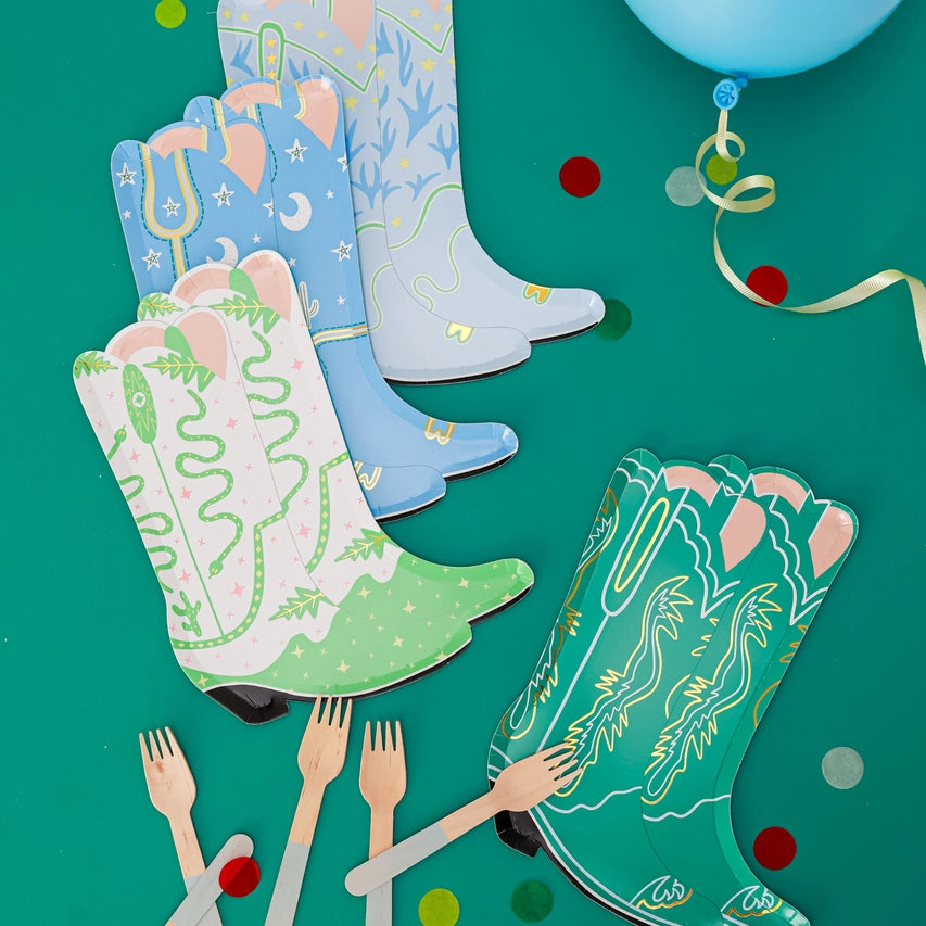 Assorted green background with four stacks of boot‑shaped plates (white‑green, pale blue, sky blue, emerald) surrounded by wooden forks and confetti.
