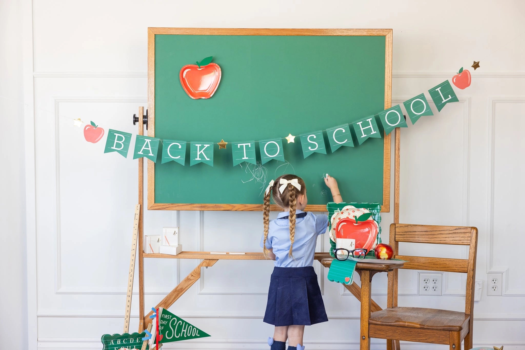 Little girl writing on a chalkboard under a Back to School Banner strung across a green board with apples and stars.