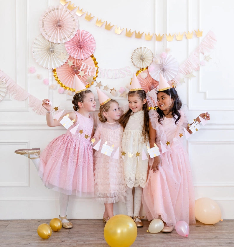 Group of four kids posing with crown-and-castle garland in front of layered pink paper fans and star banners.