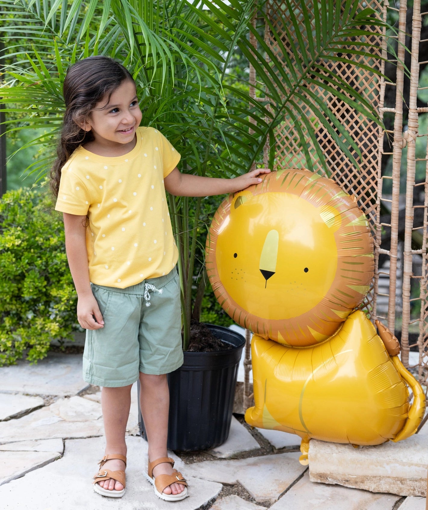 Smiling young child in a yellow shirt standing happily next to a large lion-shaped Mylar balloon at a jungle-themed birthday party.