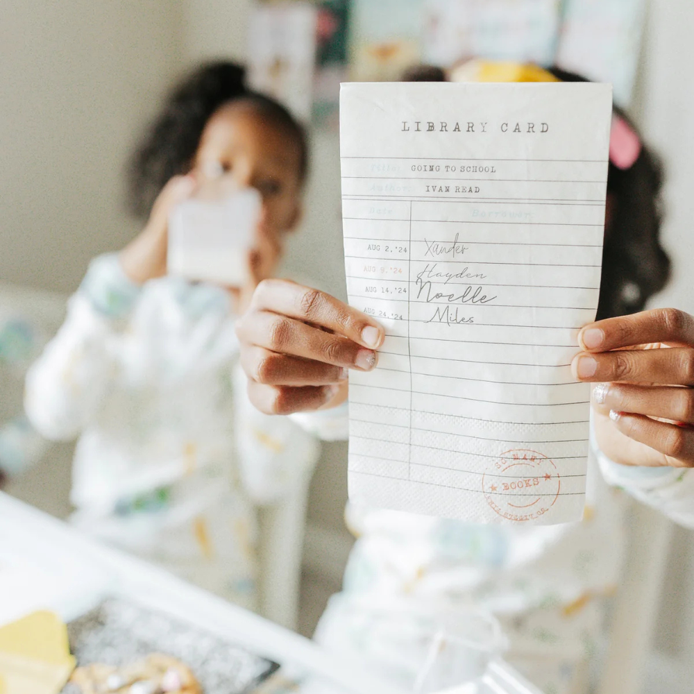 Child holding up a library card napkin with visible "borrowers" names and dates as part of a back to school celebration.