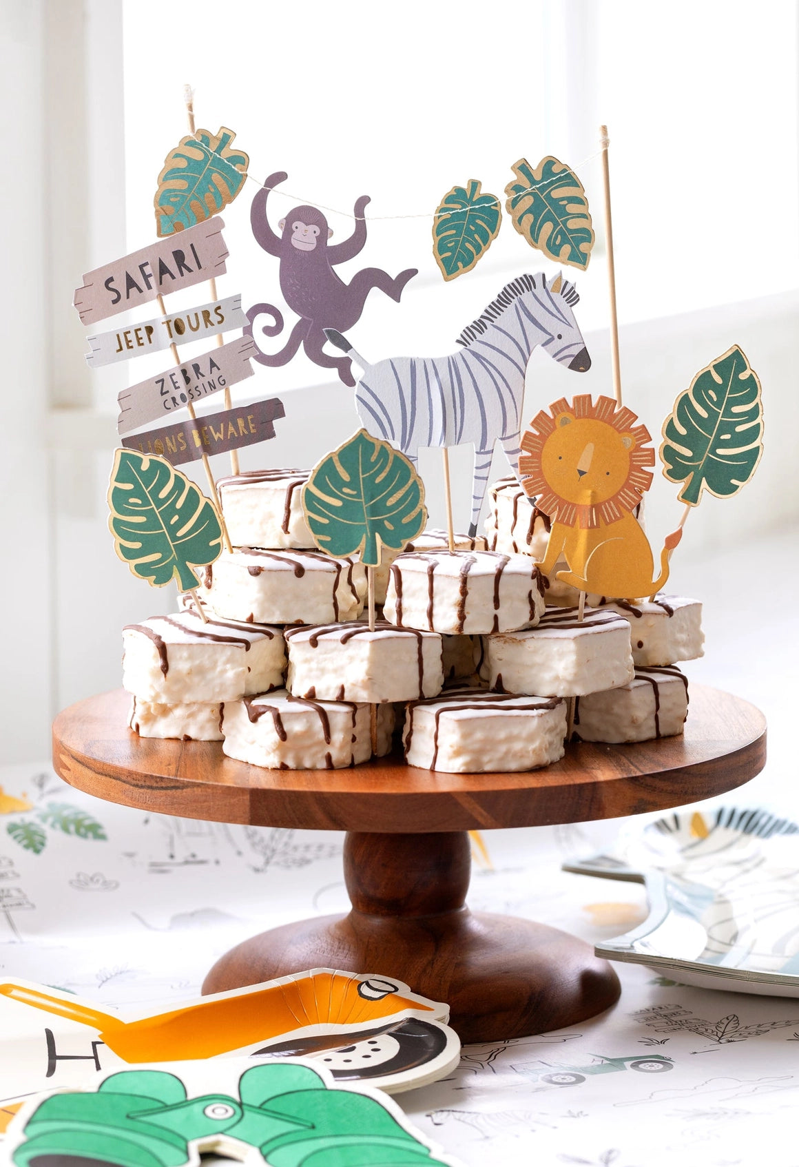 Jungle-themed cake toppers on a wooden cake stand, including zebra, lion, monkey, leaves, and safari signs.