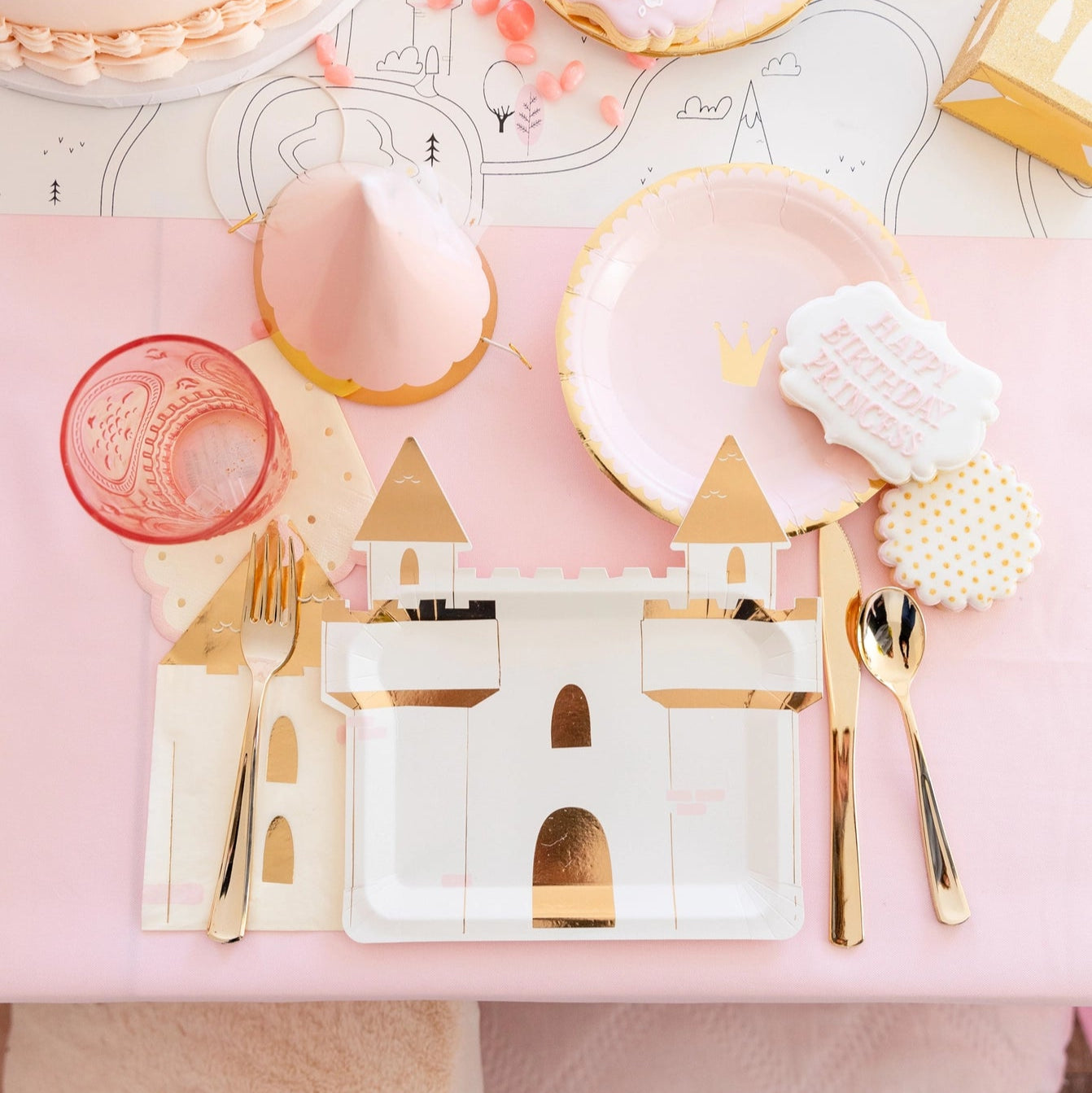 Flat-lay of birthday table featuring crown plates, cookies, and folded castle napkins alongside gold utensils.