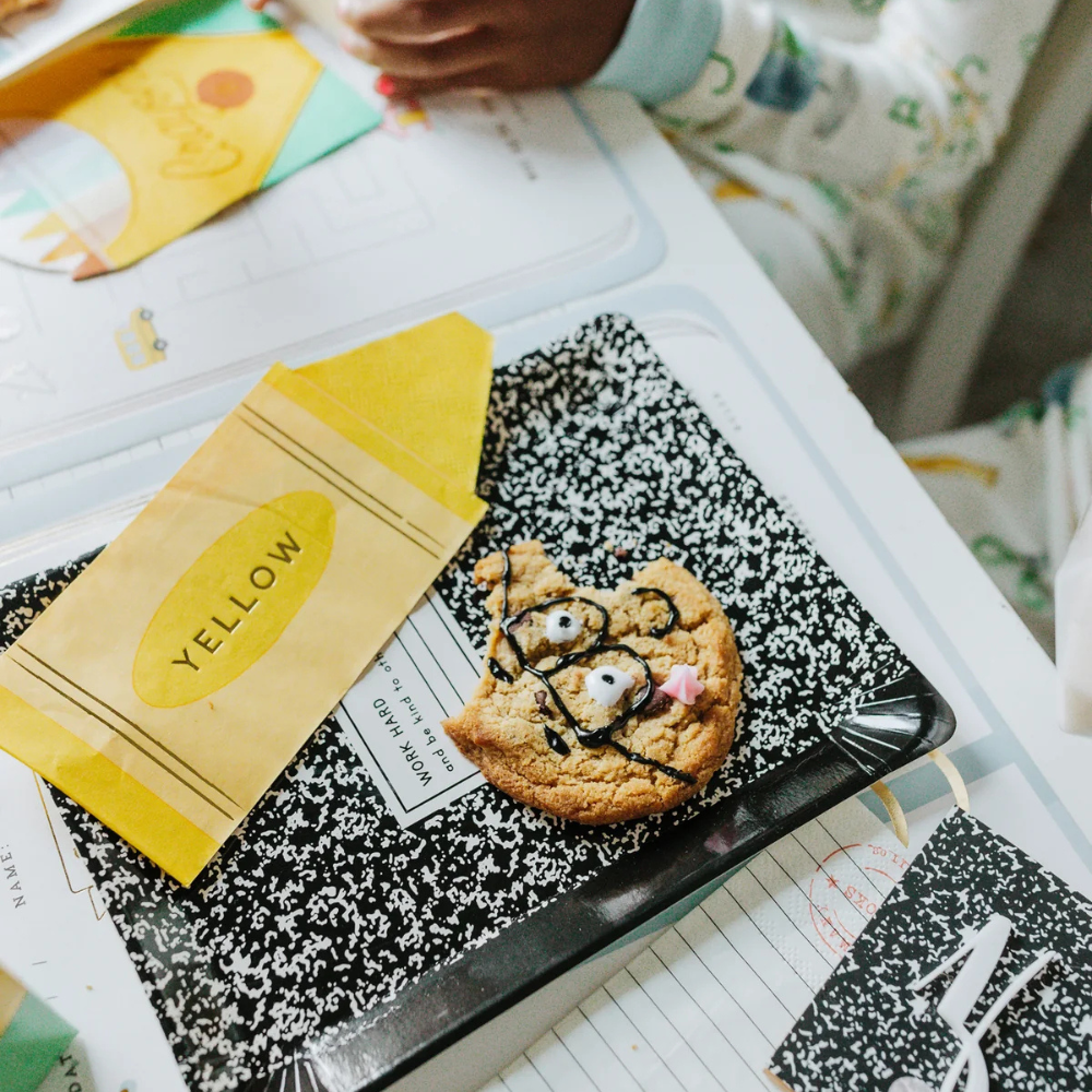 Close-up of a yellow crayon napkin placed on a black and white composition notebook next to a decorated cookie.