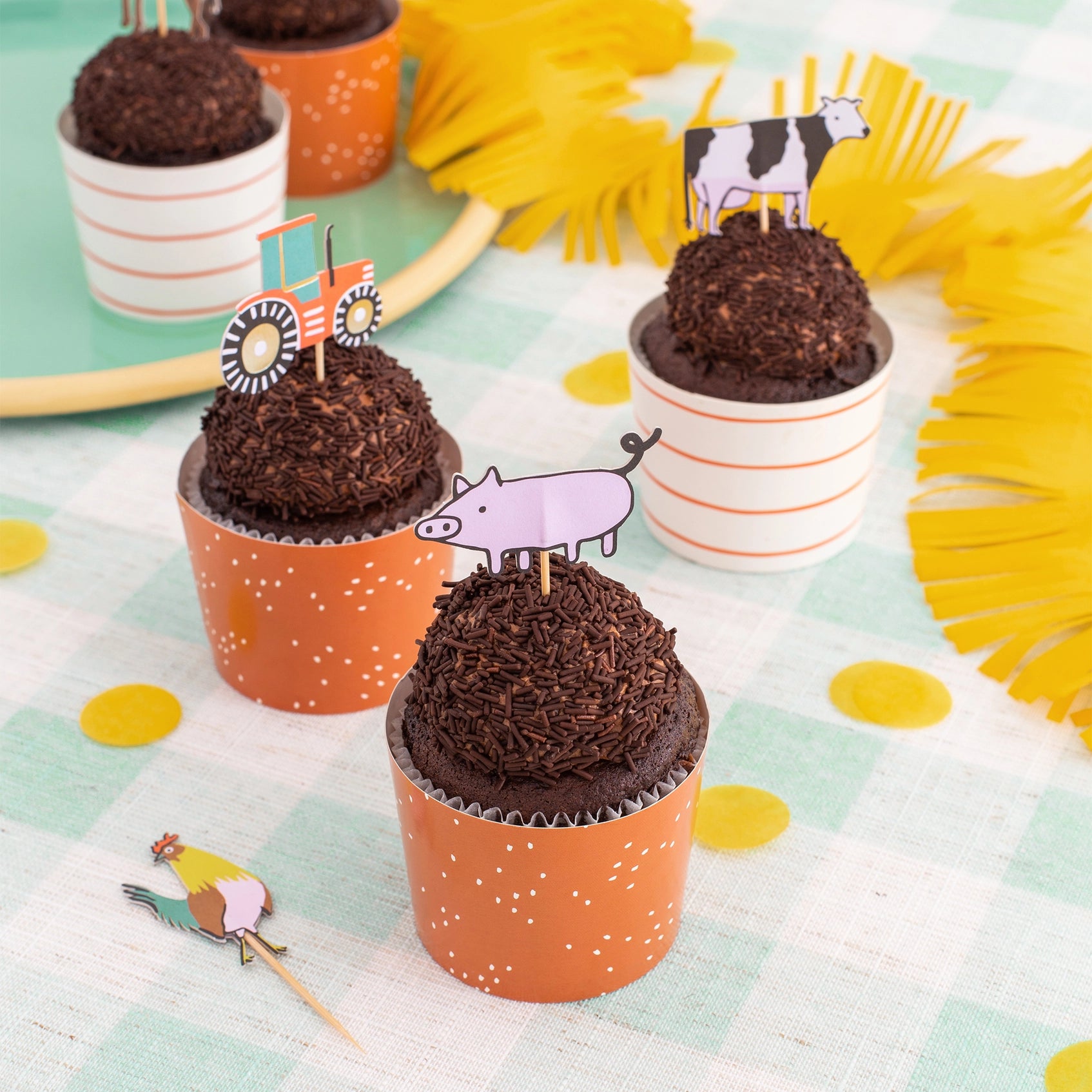 Chocolate cupcake in orange speckled cup with pig, cow, and tractor toppers on gingham table.
