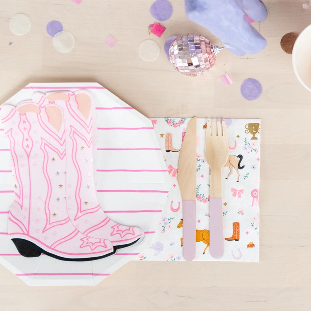 Top-down view of a light wood table set for a pony birthday: pink-striped cups with paper straws, a lavender pony figurine, pastel confetti, disco-ball globes, and four napkins—some showing pink cowgirl boots, others the horse-print pattern—paired with matching plates and utensils.