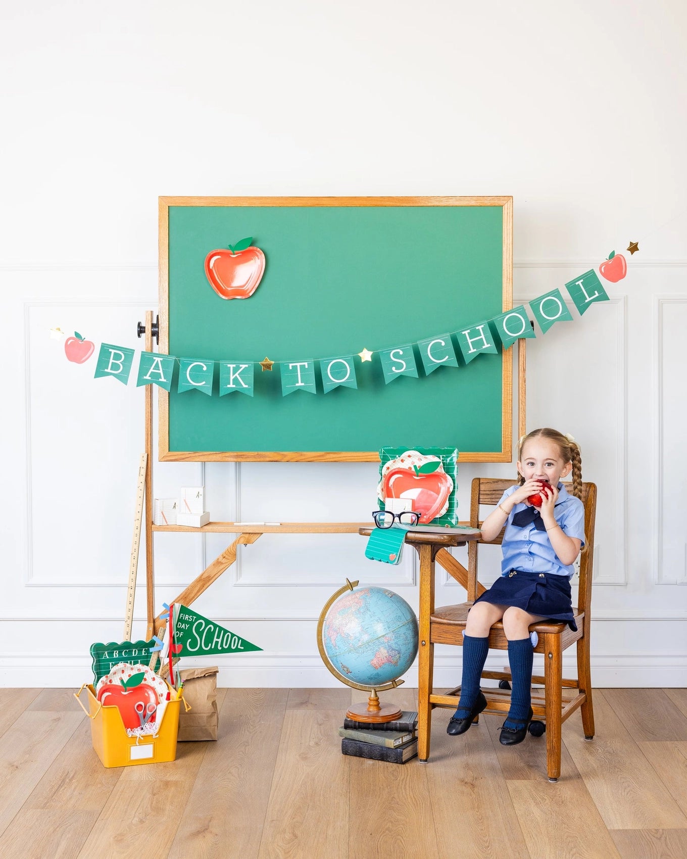 Child sitting at a vintage school desk next to a chalkboard with the banner overhead and school props below.