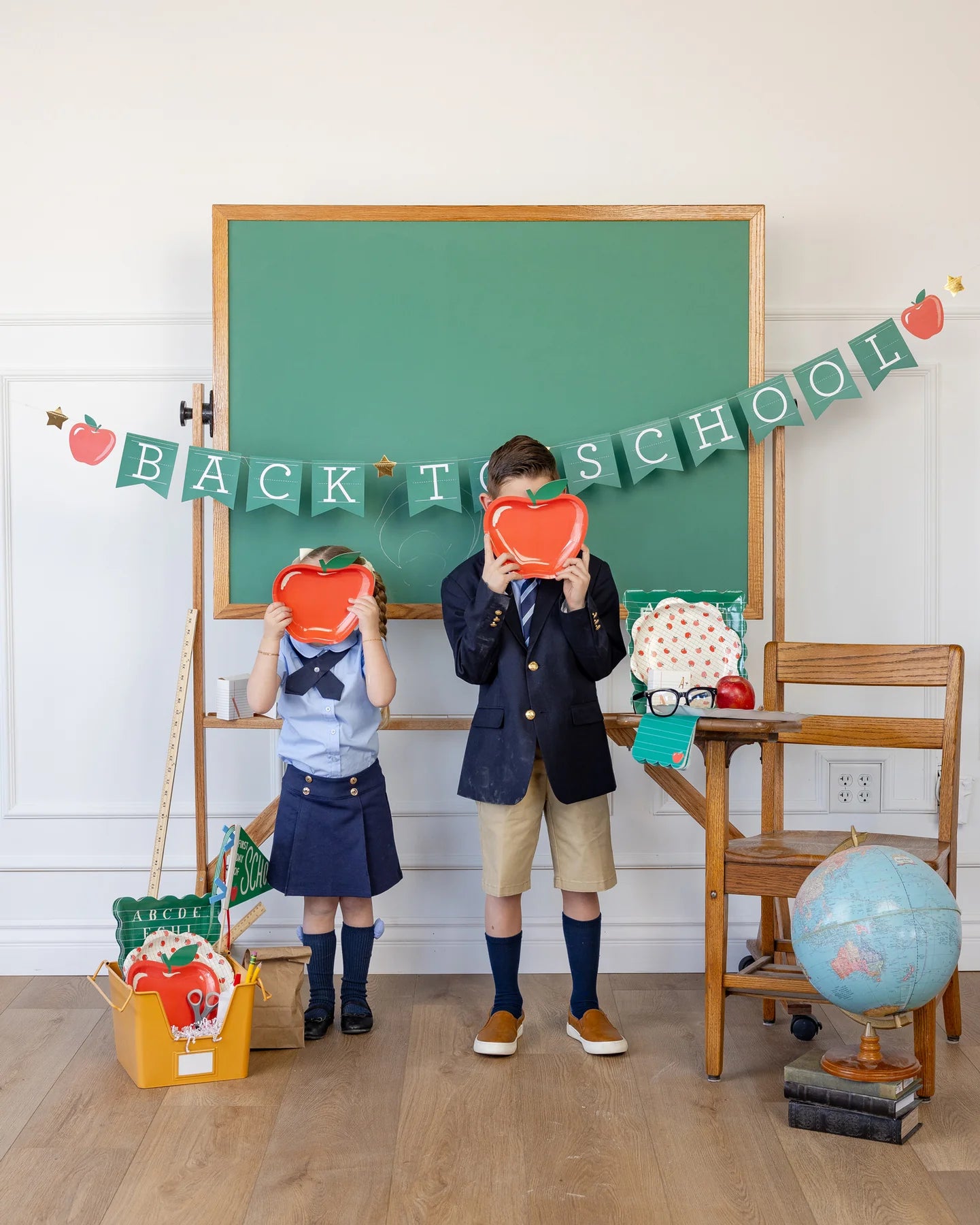Two kids standing in front of a green chalkboard holding up apple-shaped plates over their faces.