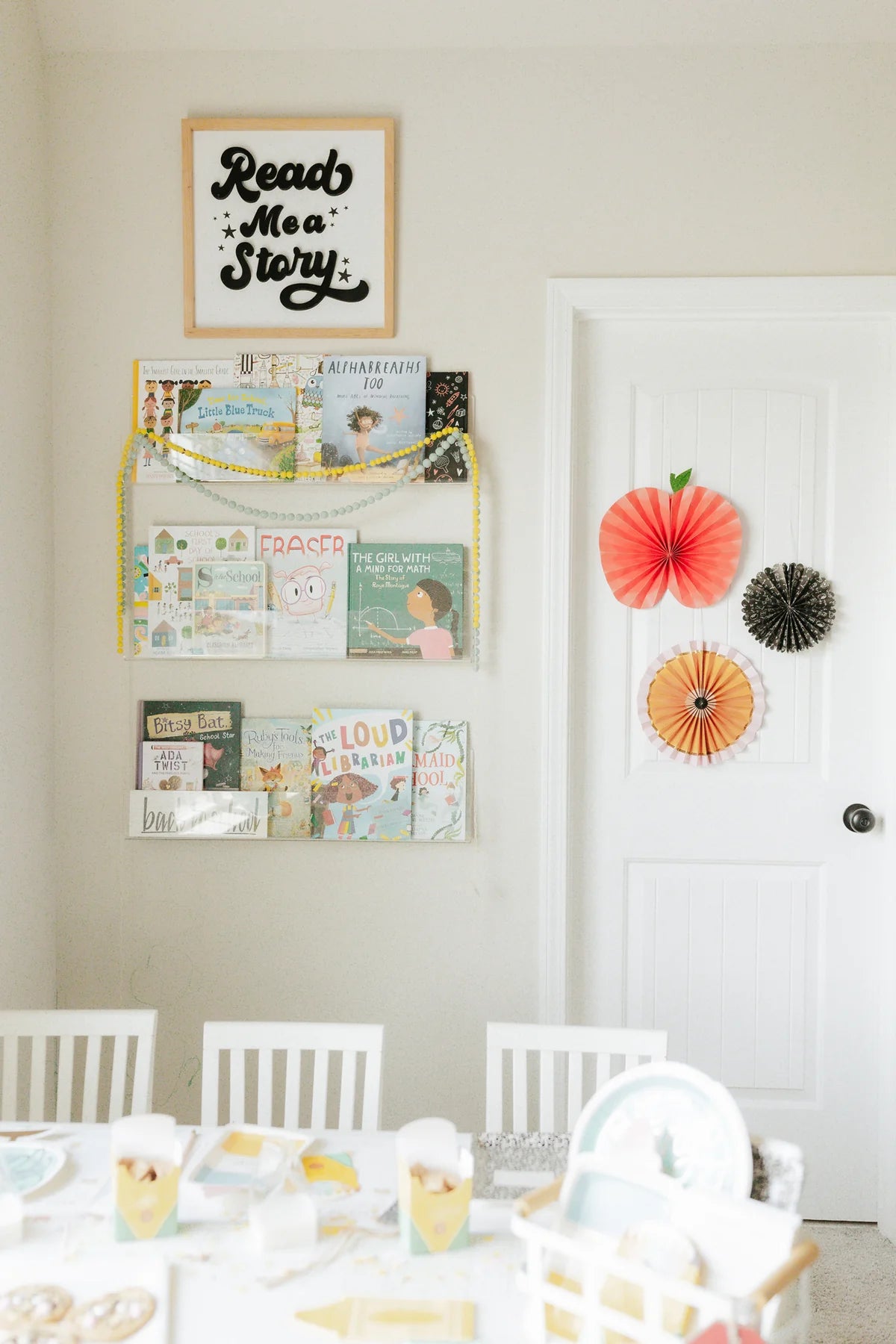  A cozy reading nook with shelves full of children’s books, yellow garland, and paper fans (apple, pencil, chalkboard) hanging on the door.